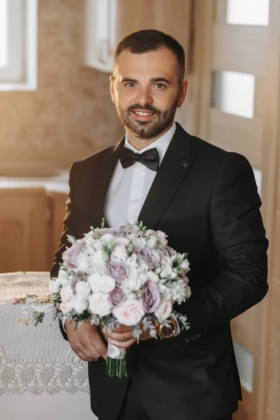 A stylish groom is preparing for the wedding ceremony in the morning. Groom\'s morning. A man in a classic suit and a bow tie. Young and handsome groom with a bouquet of flowers