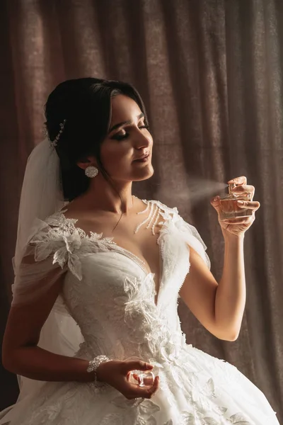 女性の美 自宅でかわいい女性 花嫁は彼女の体に香水を吹き付ける スタイリッシュな女性は白いドレスを着ています 繊細な香水をスプレーする 手の香水のスタイリッシュなガラス瓶 — ストック写真
