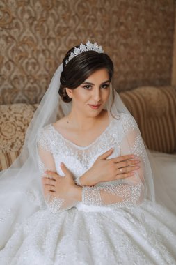 Gelin sabahları giyiniyor. Şık, gür bir elbise içinde güzel bir gelin, profesyonel saç stili, makyaj ve kafasında bir taç. Gelin kanepede oturuyor..