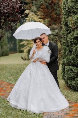 Gelin ve damat beyaz bir şemsiyeyle parkta yağmurda yürür, yağmurda düğün fotoğrafı çekilir. Muhteşem bir gelinlik içinde şemsiyesi olan bir manken. Damat gelini elinden tutar.