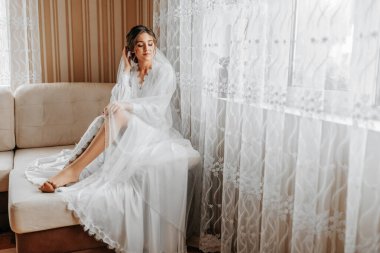 Göz kamaştırıcı genç bir gelin peçesini elinde tutarak pencerenin yanında poz veriyor. Zarif beyaz bornoz ve lüks saç stiliyle beyaz görünümlü güzel bir kadın. Düğün makyajı