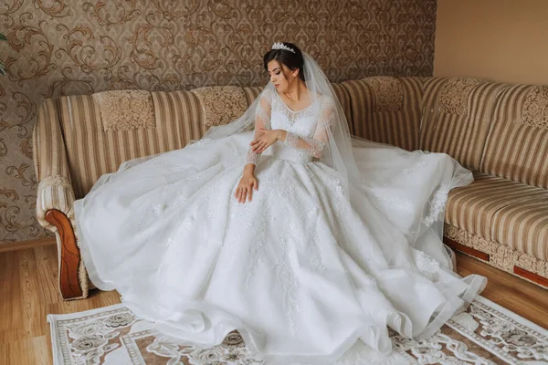 the bride gets dressed in the morning. A beautiful bride in a chic lush dress, professional hairstyle, makeup and a crown on her head. The bride is sitting in the room on the couch.