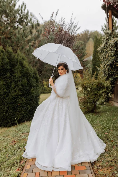 Beautiful stylish bride with a white umbrella, walks in the rain in the park, wedding photo session in the rain. A model with an umbrella in a lush wedding dress