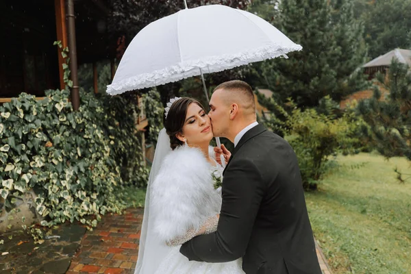 bride and groom with a white umbrella, walks in the rain in the park, wedding photo session in the rain. A model with an umbrella in a magnificent wedding dress. The groom leads the bride by the hand