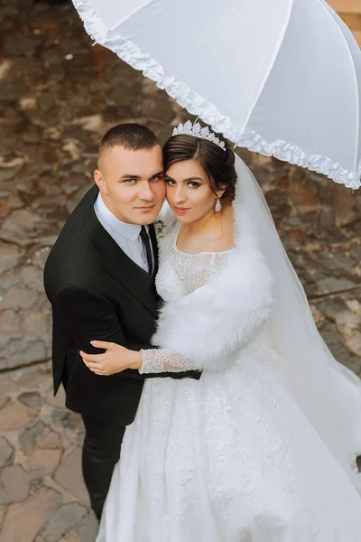 bride and groom with a white umbrella, walks in the rain in the park, wedding photo session in the rain. A model with an umbrella in a magnificent wedding dress. The groom leads the bride by the hand