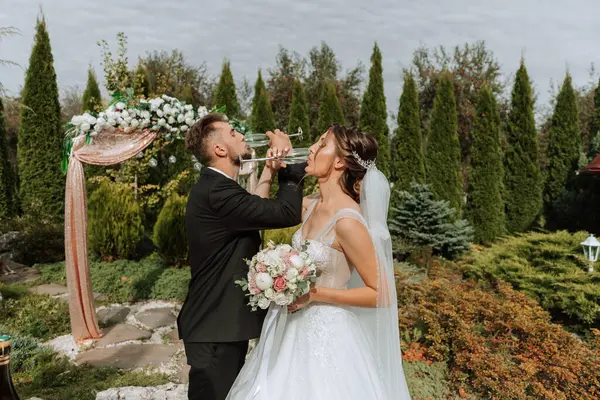 샴페인을 마시는 결혼식에서 신부와 정원의 결혼식 아치는 꽃으로 장식되어 있습니다 — 스톡 사진