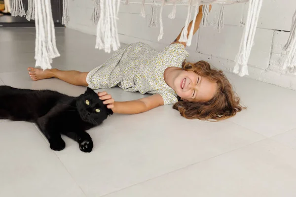 A happy child is lying on the veranda with his favorite black cat. A little girl rests on the floor of the veranda on a sunny summer day. A happy and carefree childhood.