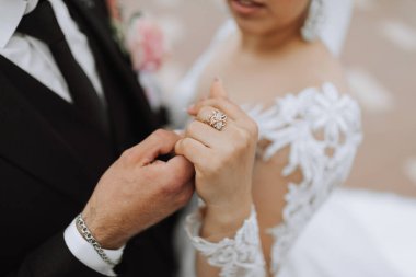 Siyah takım elbiseli şık bir damat ve uzun peçeli beyaz elbiseli şirin bir gelin. Mutlu yeni evlilerin ellerinin yakın plan düğün portresi. Bir kızın parmağında taç şeklinde bir yüzük.