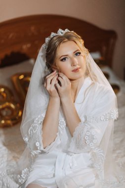 Evdeki düğün töreninden önce küpe takan genç ve güzel bir gelin. Beyaz bornozlu gelin sabah odasında küpe takıyor.