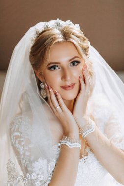 Beyaz elbiseli güzel, gülümseyen sarışın gelin yatağa oturur ve kameraya bakar. Neşeli bir kızın düğün portresi.