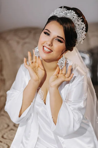 The bride in a white dress holds earrings in her hands. The bride shows off her earrings, dresses and prepares for the wedding. Professional wedding hairstyle and tiara on the head