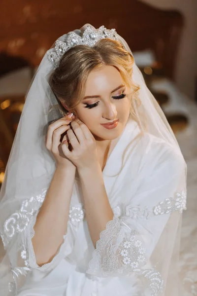 Beautiful young bride wearing earrings before wedding ceremony at home. The bride in a white robe puts on earrings in her room in the morning