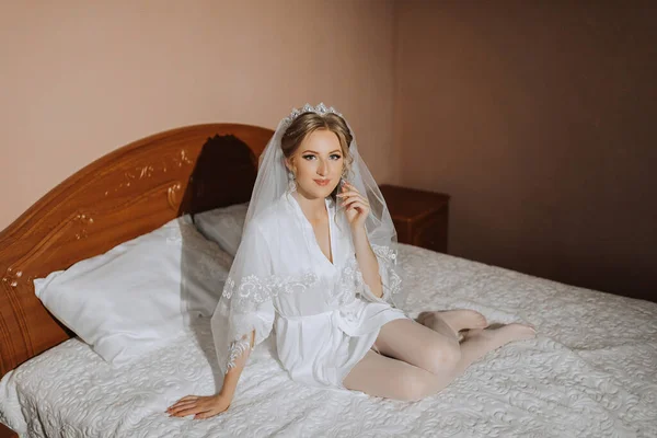 A grown-up bride sits on the bed in her room with bare legs in a white robe. Gorgeous wedding hair and makeup
