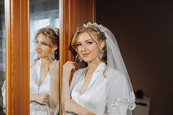Preparation for the wedding. Beautiful young bride in a white robe in the room. A luxurious model looks at her reflection in the mirror with a tiara on her head. Morning of the bride