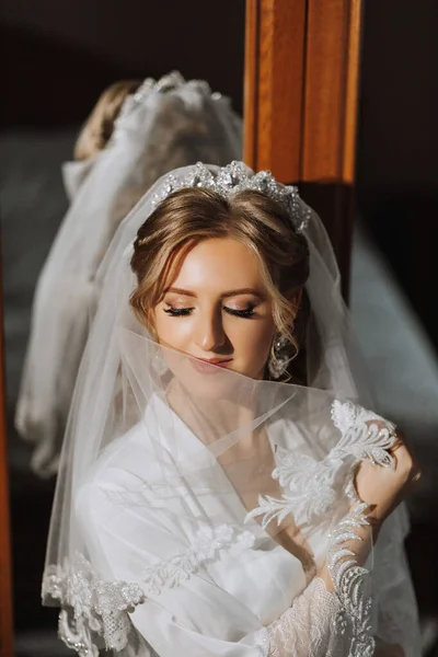 Preparation for the wedding. Beautiful young bride in a white robe in the room. A luxurious model near a mirror with a veil and a tiara on her head. Morning of the bride