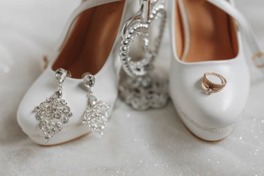Gelinin düğün ayakkabıları. Küpeleri ve altın alyansı var. Gelinliğin üstünde.