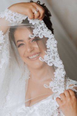 Utangaç gelin dantel elbisesinin kollarıyla kendini kapatıyor. Kafasında tacı ve profesyonel makyajı olan esmer bir gelin portresi.