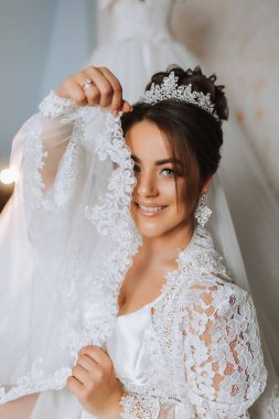 Utangaç gelin dantel elbisesinin kollarıyla kendini kapatıyor. Kafasında tacı ve profesyonel makyajı olan esmer bir gelin portresi.