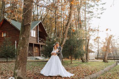 Muhteşem romantik bir yeni evli çift gün batımında çimenlerin üzerinde ağaçların yapraklarıyla bir sonbahar parkında kucaklaşıyor. Geniş açı fotoğrafı