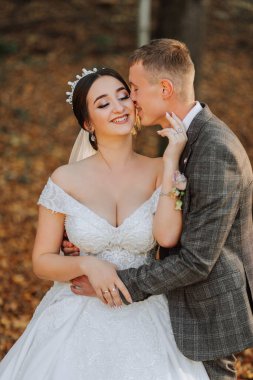 Muhteşem romantik bir yeni evli çift gün batımında çimenlerin üzerinde ağaçların yapraklarıyla bir sonbahar parkında sarılıyor.