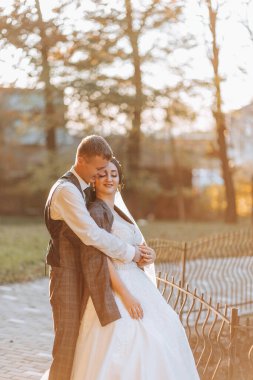Muhteşem romantik bir yeni evli çift gün batımında çimenlerin üzerinde ağaçların yapraklarıyla bir sonbahar parkında sarılıyor.