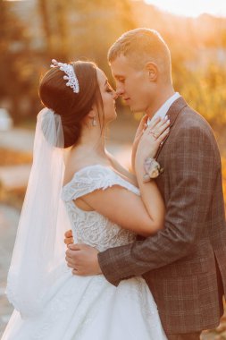 Muhteşem romantik bir yeni evli çift gün batımında çimenlerin üzerinde ağaçların yapraklarıyla bir sonbahar parkında sarılıyor.