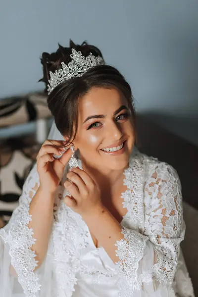 自宅での結婚式の前にイヤリングを着用する美しい若い花嫁 白いローブの花嫁は朝の部屋にイヤリングを置く — ストック写真