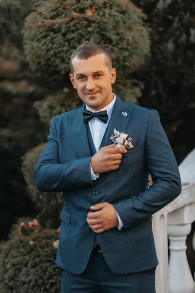 A handsome groom in a black suit and black tie is standing outdoors. Wedding portrait. A man in a classic suit against the background of autumn leaves