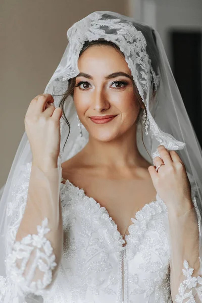 Close-up shot of an elegant brunette bride in a white dress posing under a veil close-up. Bride portrait, professional wedding makeup and hairstyle, bridal fashion. Beautiful bride in a veil