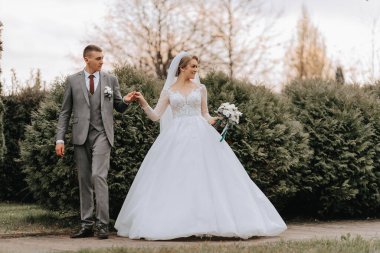 Damat ve gelin parkta uzun yeşil ağaçların arasında el ele tutuşuyorlar. Geniş açılı sonbahar fotoğrafı. Gönüllü gelinlik. Damadın klasik takımı.