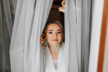 Sabahları beyaz pijamalı güzel ve nazik gelin balkondaki pencereden bakıyor. Güzel doğal makyaj. Düğün saç stili. Gelinin düğün hazırlıkları..