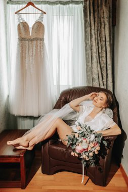 Pencere kenarında beyaz pijamalı gelinin portresi elinde bir buket çiçekle bir sandalyede oturuyor. Güzel doğal makyaj. Düğün saç stili. Gelinin sabahı.