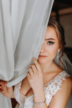 Gelinlik giymiş güzel ve hassas bir gelin, sabahları pencerenin yanında durur ve yüzünün yarısını duvakla kapatır. Güzel doğal makyaj. Düğün saç stili. Gelinin sabahı.