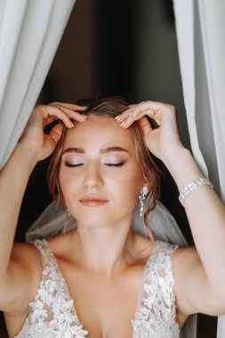 Gelinlik giymiş güzel ve hayalperest bir gelinin portresi, sabah, pencerenin önünde poz verirken. Güzel doğal makyaj. Düğün saç stili. Gelinin sabahı.