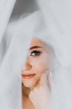 Gelinlik giymiş güzel ve hassas bir gelin, sabahları pencerenin yanında durur ve yüzünün yarısını duvakla kapatır. Güzel doğal makyaj. Düğün saç stili. Gelinin sabahı.