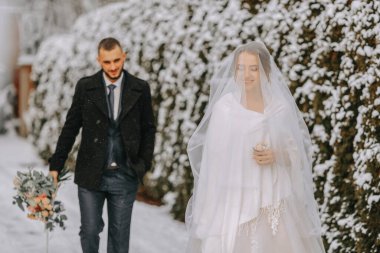 Neşeli, güzel yeni evlilerin hassas portresi. Damat kış parkındaki büyüleyici gelinine doğru yürür. Gelinlik ve pançolu gelin. Damat siyah bir ceket giyiyor.
