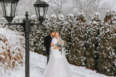 Mutlu yeni evlilerin hassas portresi. Damat kış parkında geline sarılır. Gelinlik ve beyaz pançolu gelin. Damat siyah bir ceket giyiyor. Geniş açı. Boş alan