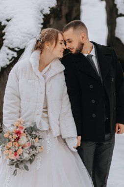 Kışın düğün. Beyaz vizon kürklü, elinde bir buket çiçekle şık bir gelin ve soğuk havada beyaz karlı bir ormanda siyah ceketli bir damat..