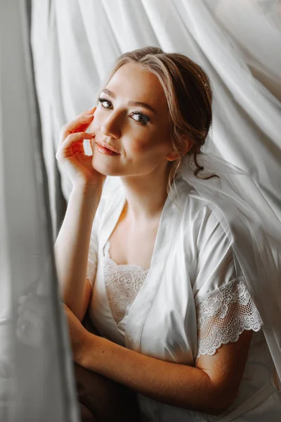 beautiful and gentle bride in white pajamas in the morning, posing near the window with natural light. Beautiful natural makeup. Wedding hairstyle. Morning of the bride.