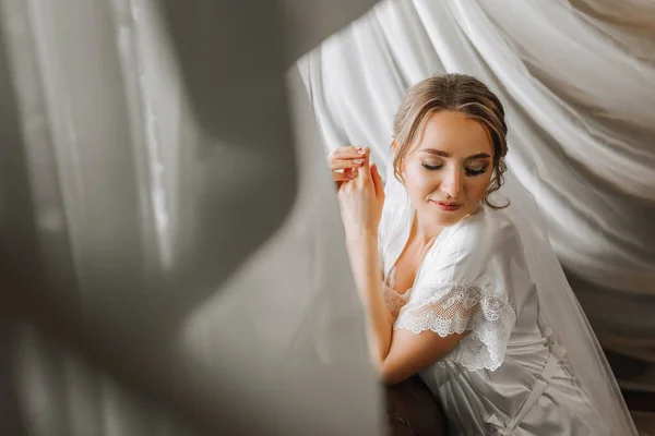 beautiful and gentle bride in white pajamas in the morning posing near the window with natural light. Beautiful natural makeup. Wedding hairstyle. Morning of the bride. Horizontal photo. Free space