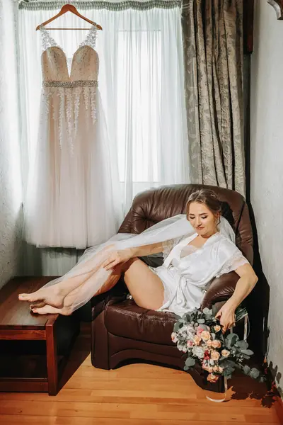 a beautiful and gentle bride in white pajamas is posing by the window, sitting on a chair with a bouquet of flowers in her hands. Beautiful natural makeup. Wedding hairstyle. Morning of the bride.
