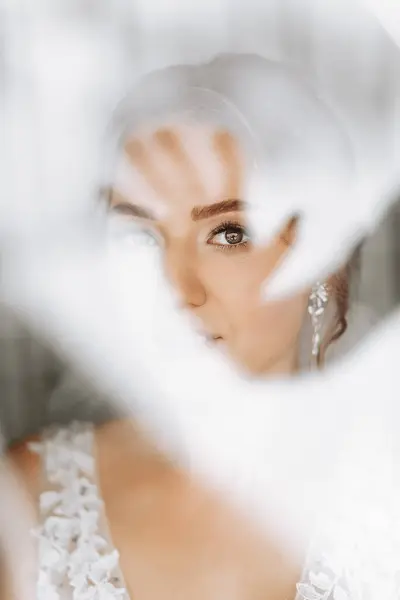 a beautiful and gentle bride looks out the window in the morning. Photo through the window. Beautiful natural makeup. Wedding hairstyle. Morning of the bride.