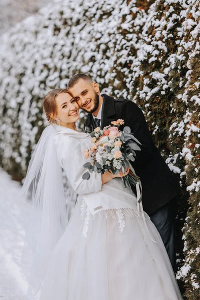 雪に覆われた木の背景にある幸せな新生児の肖像画 ウィンターパークで花嫁を抱きしめる ウェディングドレスとホワイトポンチョの笑顔の花嫁 グロムは黒いコートを着ています — ストック写真