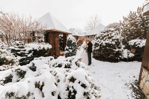 happy newlyweds between snowy trees. The groom hugs the bride in the winter park. Smiling bride with a bouquet of flowers in a wedding dress and white poncho. The groom is dressed in a black coat.