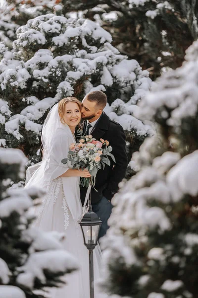 Portrait of the bride and groom between snowy trees. The groom hugs the bride in the winter park. Bride with a bouquet of flowers in a wedding dress and poncho. The groom is dressed in a black coat.