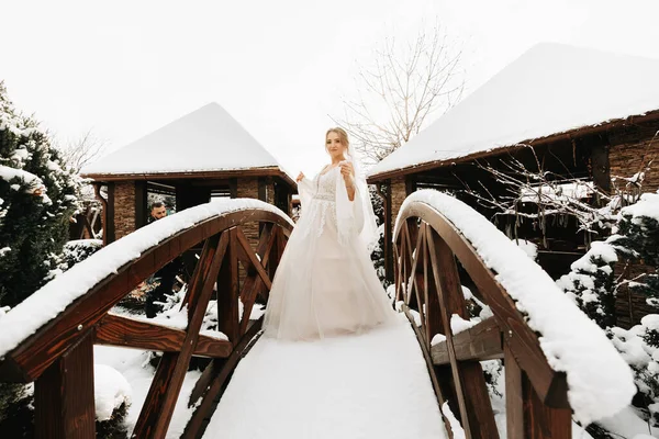 Bride in snow on wooden bridge, portrait of beautiful bride in white poncho and wedding dress in winter park