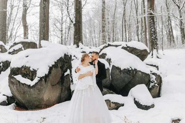 冬の結婚式について 花嫁と新郎は雪に覆われた岩の背景に立っています 白いドレスと白いポンチョの花嫁 黒いコートで成長する — ストック写真