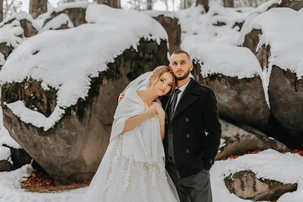 Wedding in winter. The bride and groom are standing against the background of snow-covered rocks. The bride in a white dress and white poncho. Groom in a black coat