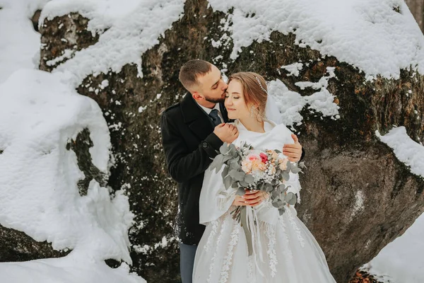 Wedding in winter. Stylish bride in white poncho and groom in black coat in forest with white snow in cold weather. The groom kisses the bride against the background of snowy rocks