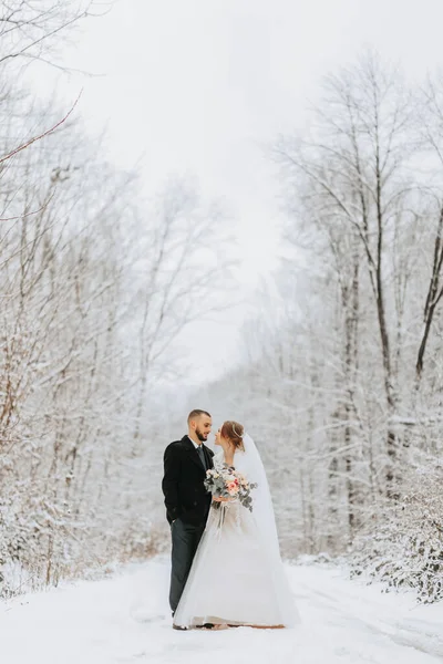 Beautiful wedding couple standing in winter snowy forest, woman in white dress and mink fur coat, bearded man in black coat